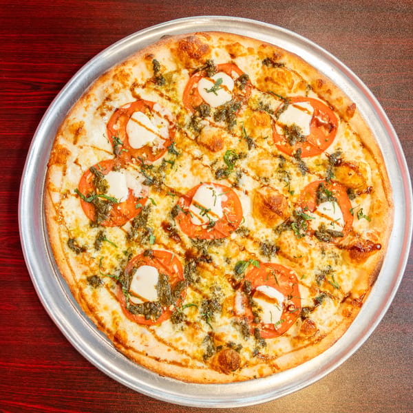 A whole round pizza with mozzarella, sliced tomatoes, pesto, and balsamic glaze on a metal tray.