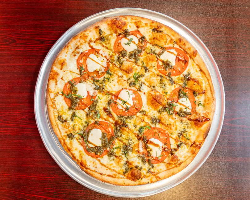 A whole round pizza with mozzarella, sliced tomatoes, pesto, and balsamic glaze on a metal tray.