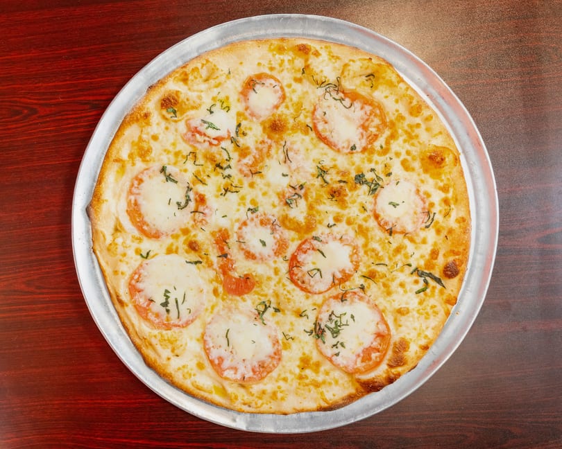 A whole Margherita pizza with fresh mozzarella, sliced tomatoes, and basil on a metal tray.